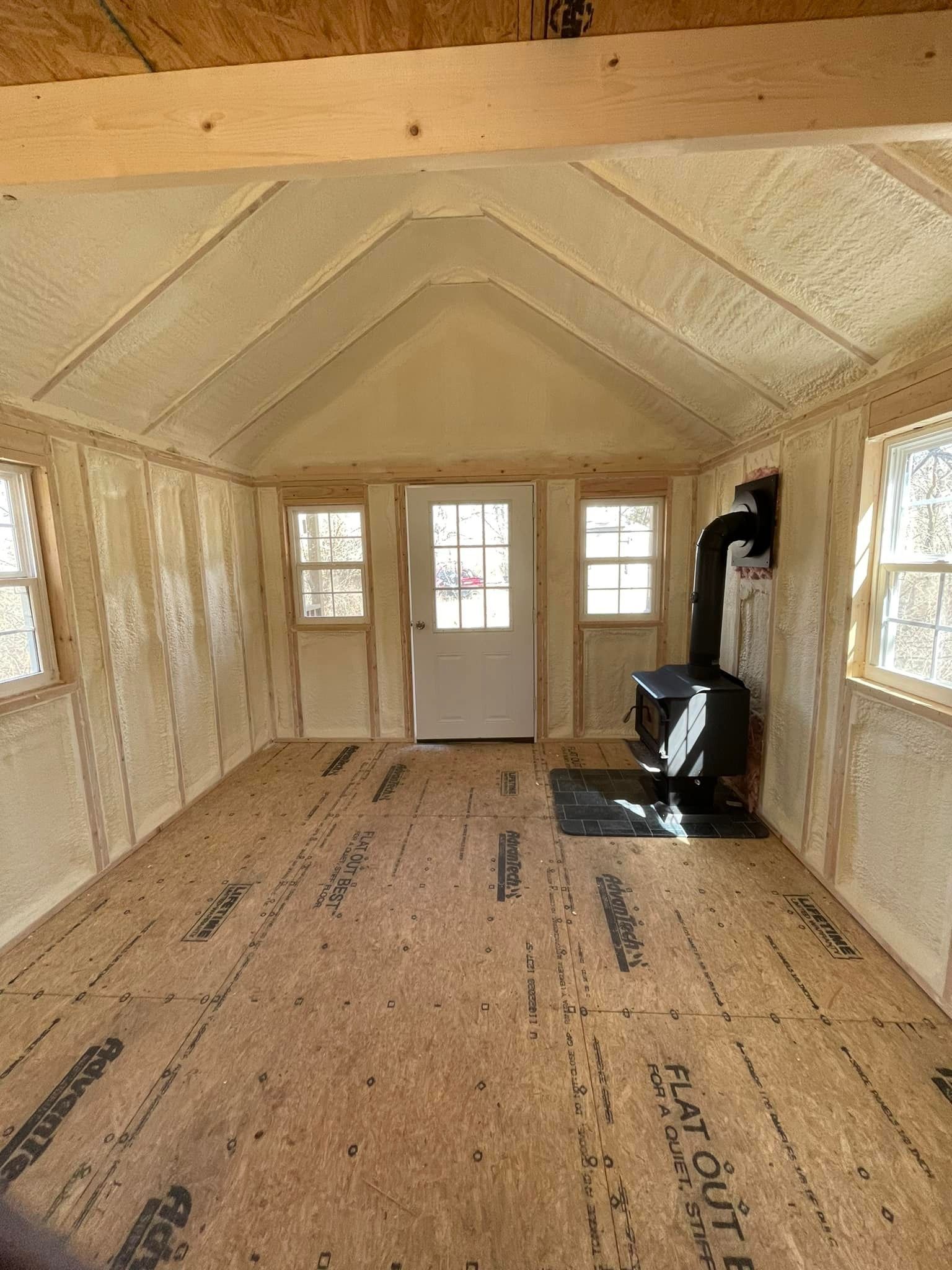 Insulated tiny house interior with plywood floor, white door, windows, and a wood stove at one end
